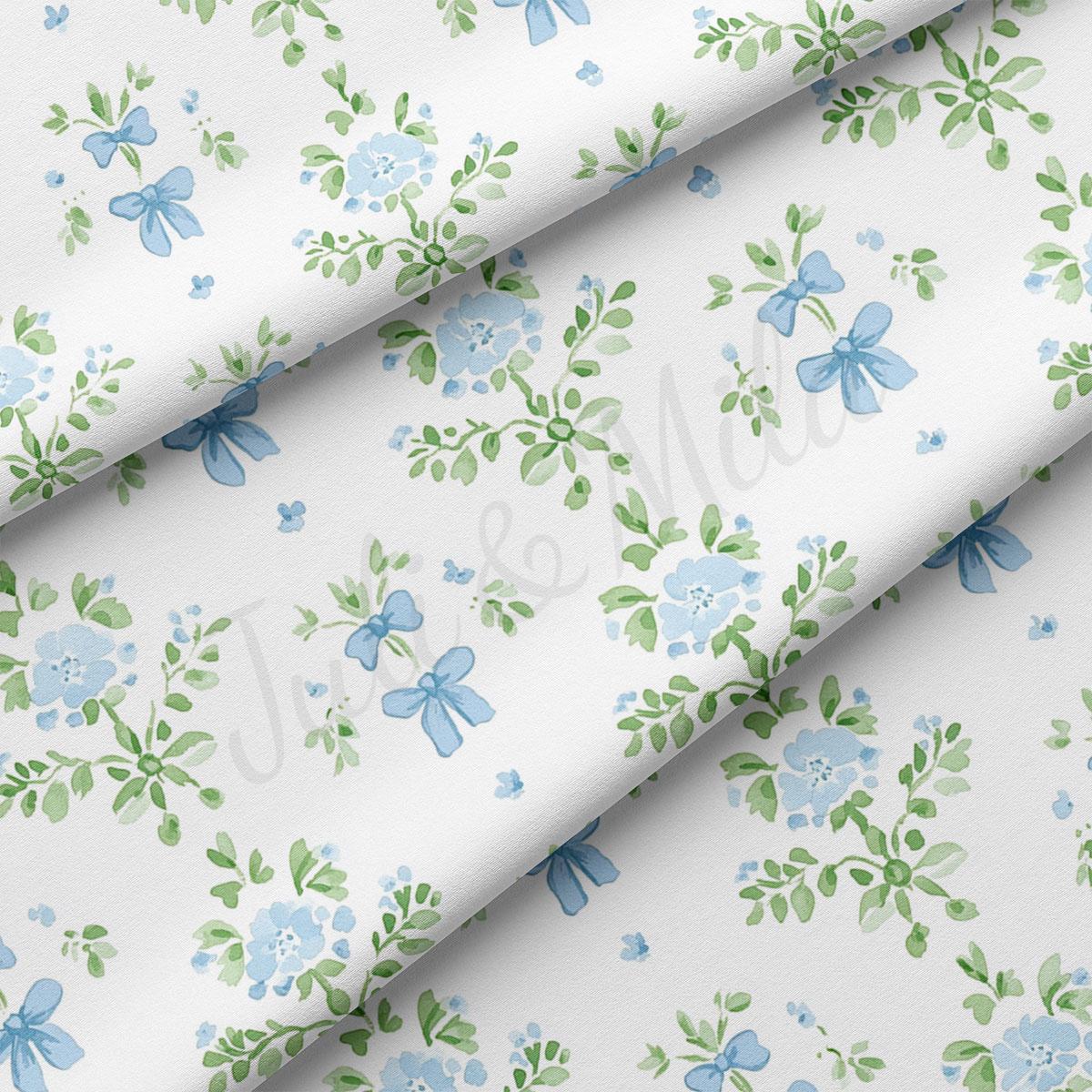 a close-up view of a fabric with a repeating floral pattern in shades of blue and green. The pattern consists of various flowers and leaves, some of which are adorned with blue bows. The fabric appears to be white, providing a contrast to the vibrant colors of the floral design. The pattern repeats across the fabric, creating a sense of continuity and rhythm.